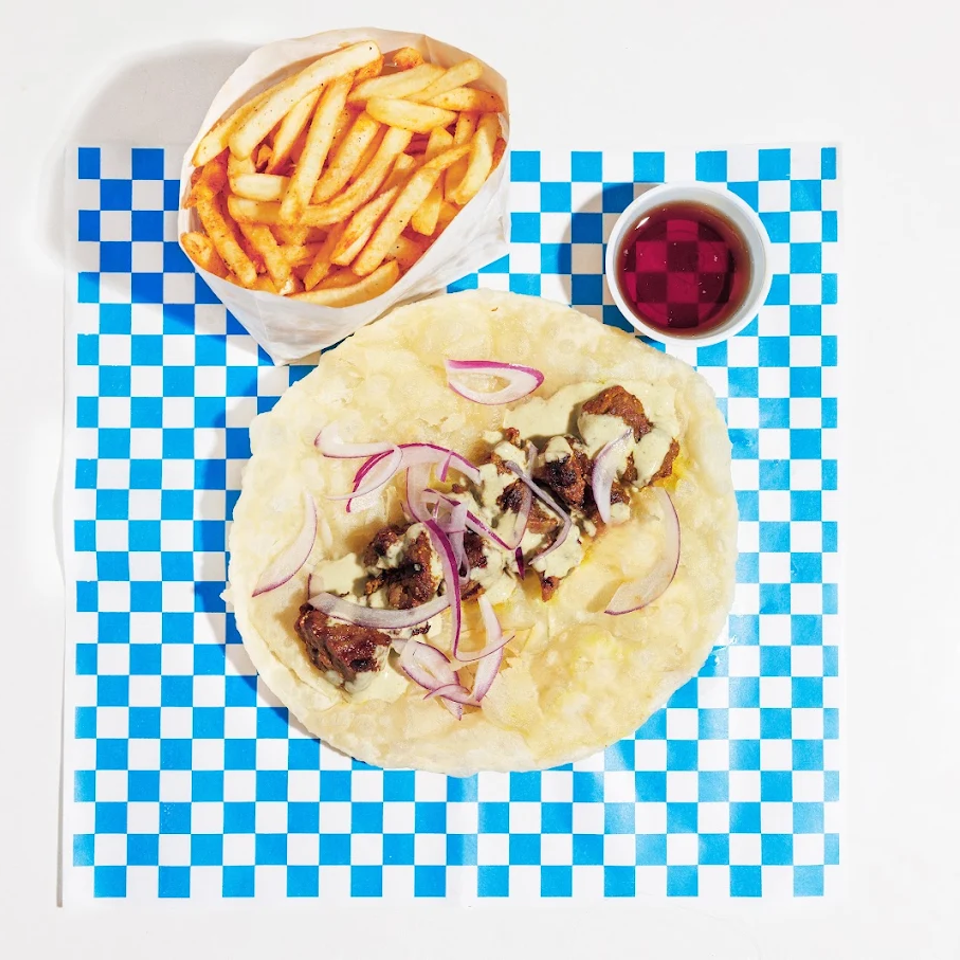 Overhead view of a flatbread topped with grilled meat, creamy sauce, and red onion, served with fries and dipping sauce.